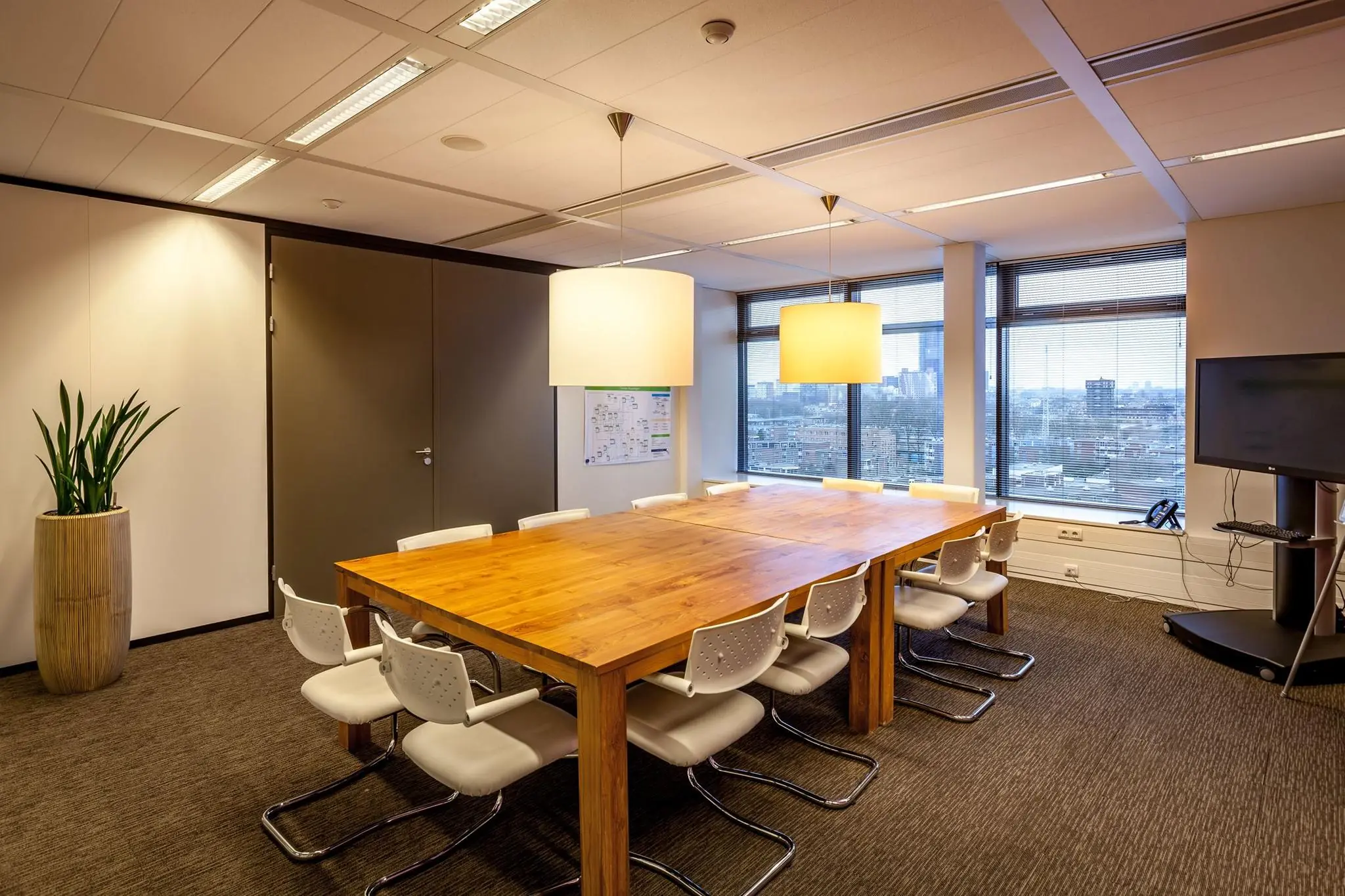 Vergaderruimte met een grote houten tafel, witte stoelen, hanglampen en uitzicht op de stad aan de Prinses Beatrixlaan.